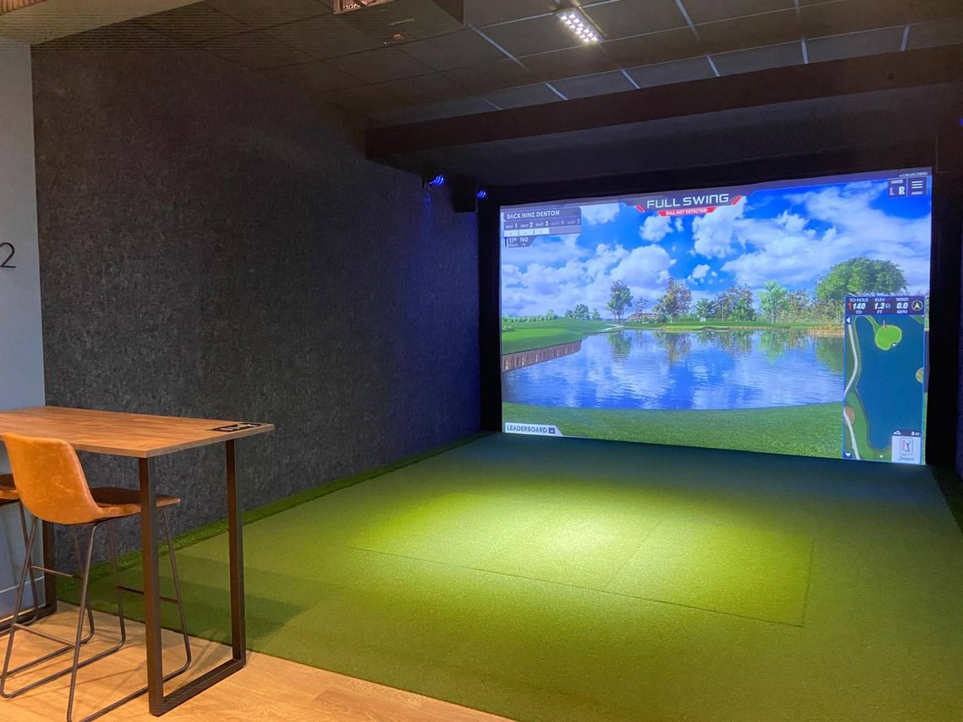 A finished golf simulator bay at The Back Nine Golf in Denton, TX, with the screen lit, a putting surface and a high-top table